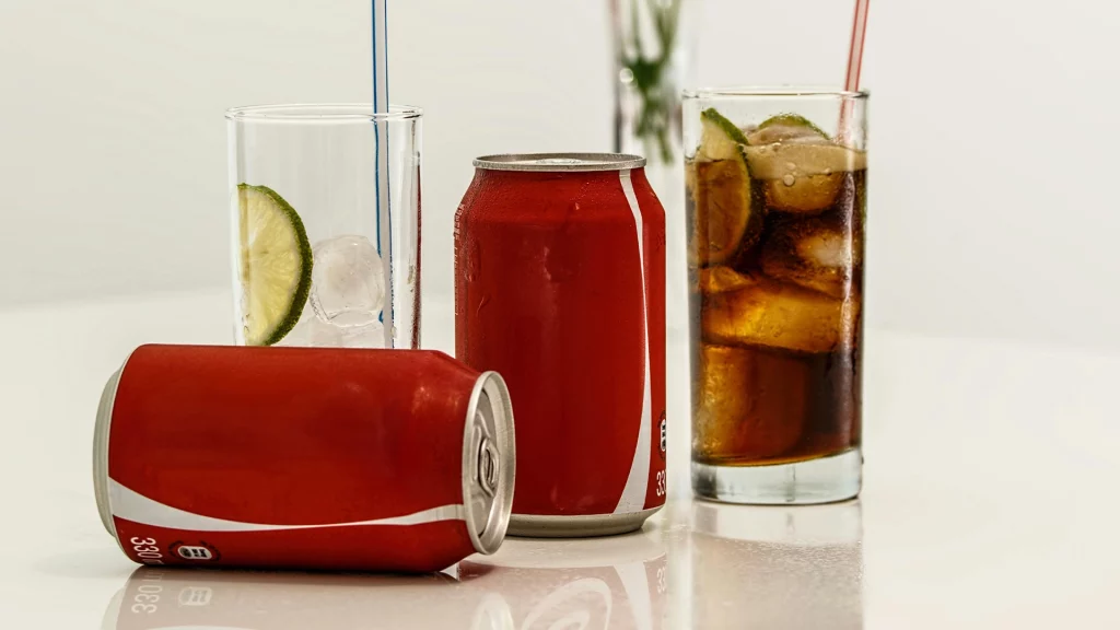 Coca Cola Dosen mit zwei Gläsern. Eine Dose liegt auf der Seite, eine steht. Das eine Glas ist nur mit Eiswürfeln gefüllt, das andere ist zudem mit Cola gefüllt. Auf den Cola Dosen fehlt das Logo